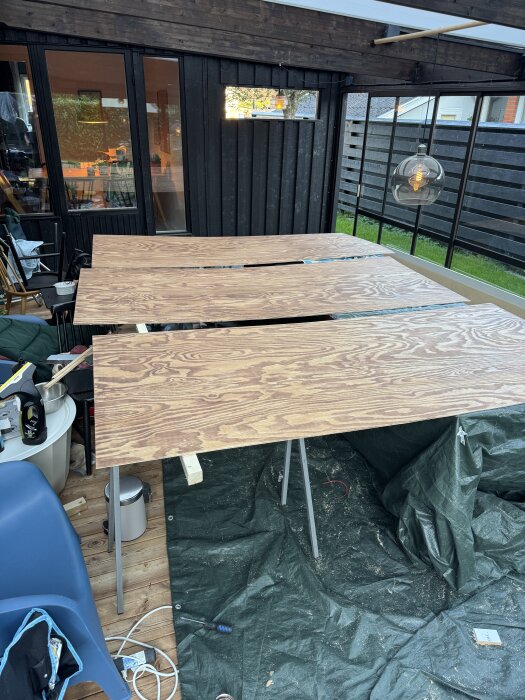 Tre plywoodskivor ligger på bockar i ett uterum, omgivna av verktyg och en hängande lampa i bakgrunden.