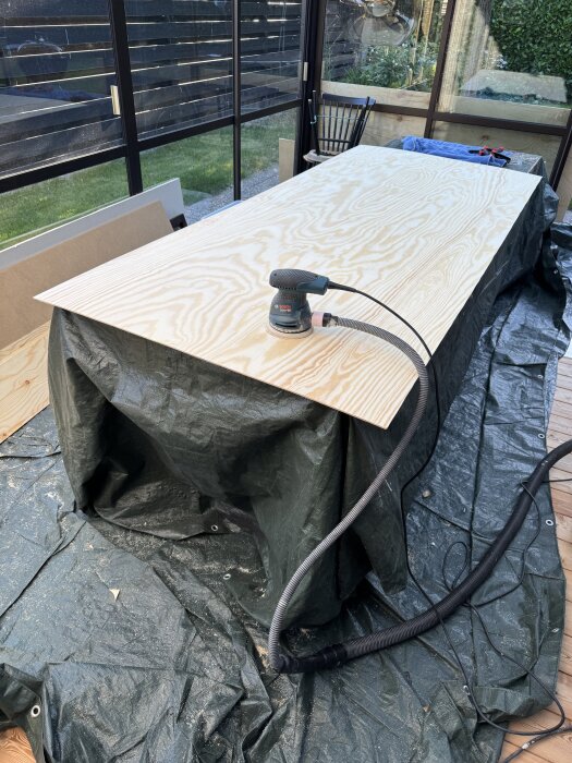 Plywoodskiva på ett bord i uterummet, slipas med en elektrisk slipmaskin; en presenning ligger under.