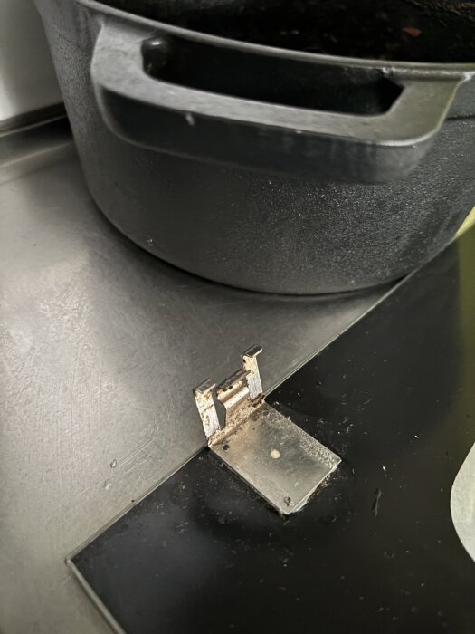 Närbild på en metallfäste med tre pinnar på en svart yta bredvid en gryta, del av ett ugnsluckemonteringsproblem.