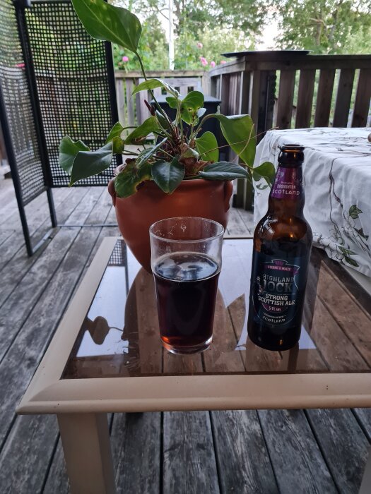 Flaska med Highland Jock Strong Scottish Ale, ett glas öl och en krukväxt på ett glasbord på en uteplats.