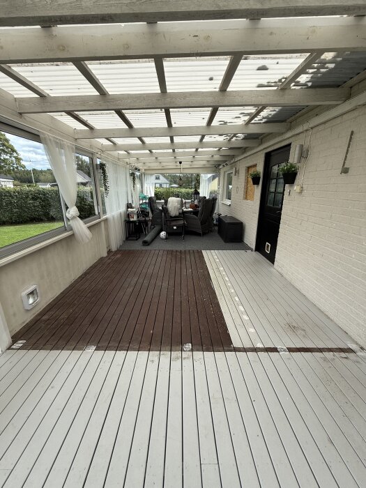 Målat altangolv med grå och brun yta, skjutpartier flyttade. Uteplats med möbler, vita väggar och tak, inramad av grönska och trädgård.