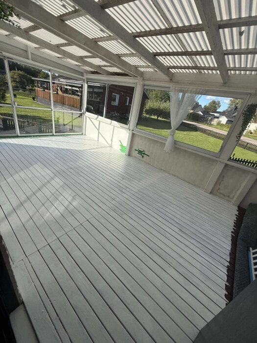 Nymålat golv på inglasad altan med utsikt mot grönskande trädgård, förenad yta utan synlig uppdelning mellan ute- och innedel.