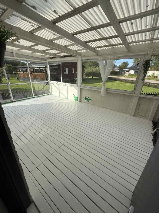 Nymålat golv på en inglasad altan, skjutpartier och gardiner synliga, ger en enhetlig och sammanhängande yta mot trädgården.