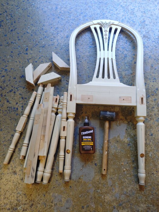 Delar av en stol inkl. ryggstöd, trästycken, lim och hammare, liggande på ett golv.