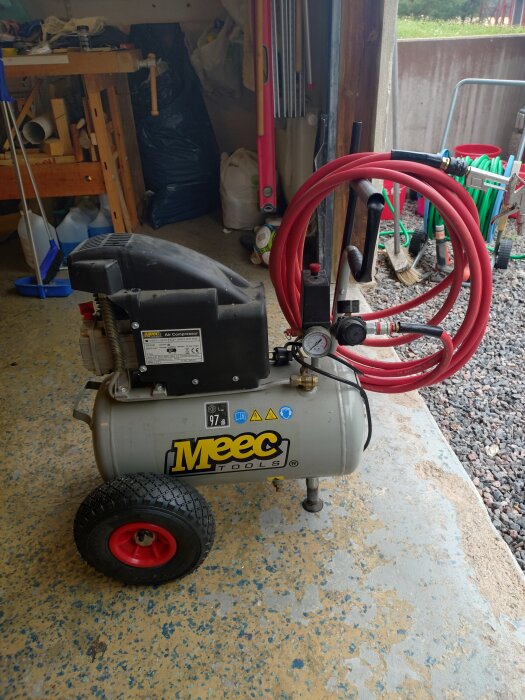 Kompakt kompressor med rödmålade hjul och uppgraderat handtag, röd slang som hållare, placerad på ett verkstadsgolv bredvid ett grusområde.