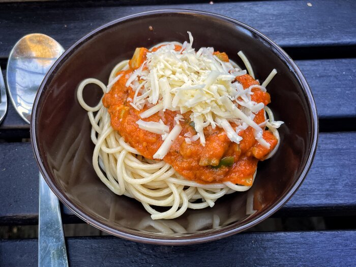 Skål med spaghetti toppad med tomatsås med grönsaker och riven ost på ett bord, bredvid en sked.