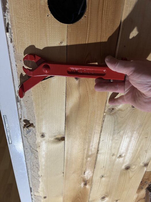 Hand håller i en röd brytjärn mot en trävägg med slitet trä och ett runt hål.