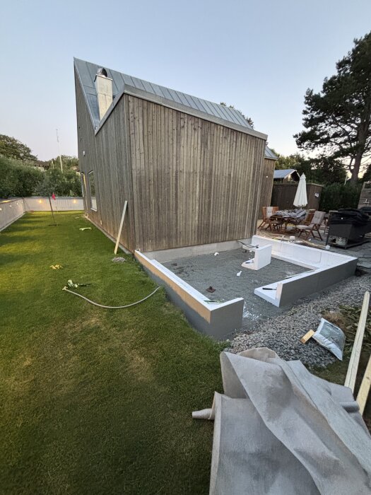 Kantelement installerade på en gjuten yta vid en träbyggnad; gräsmatta och stenläggning omkring, byggmaterial syns också i bilden.