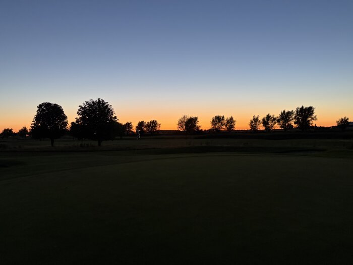 Solnedgång över en golfbana med silhuetter av träd vid horisonten och en flagga på greenen under en klar kvällshimmel.