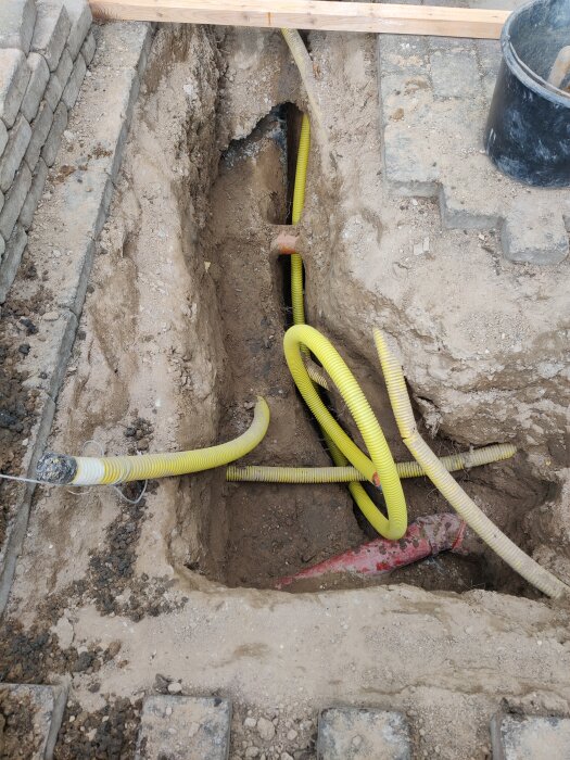 En uppgrävd schaktdike med synliga gula skyddsrör, en rödledning och löst jord. Stenläggning och ett svart plasthink syns vid kanten.
