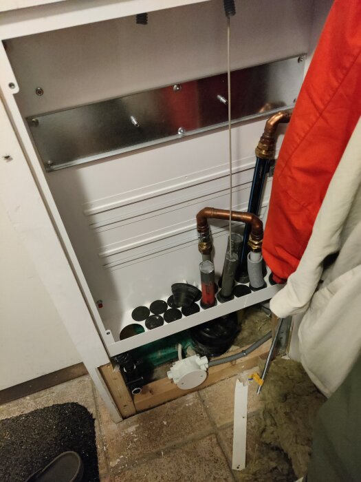 Ett skåp med vattenmätare och kopplingar, rörböjar och olika rör syns. Rören är ordnat och anslutna i skåpet mot en bakgrund av vit panel.