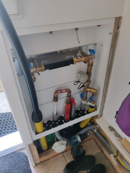 Vattenmätarskåp med ny rörinstallation. Inkommande vattenledning, kopparrörsloop för varmvatten, backventil samt vattenfelsbrytare synliga. Verktyg på golvet.