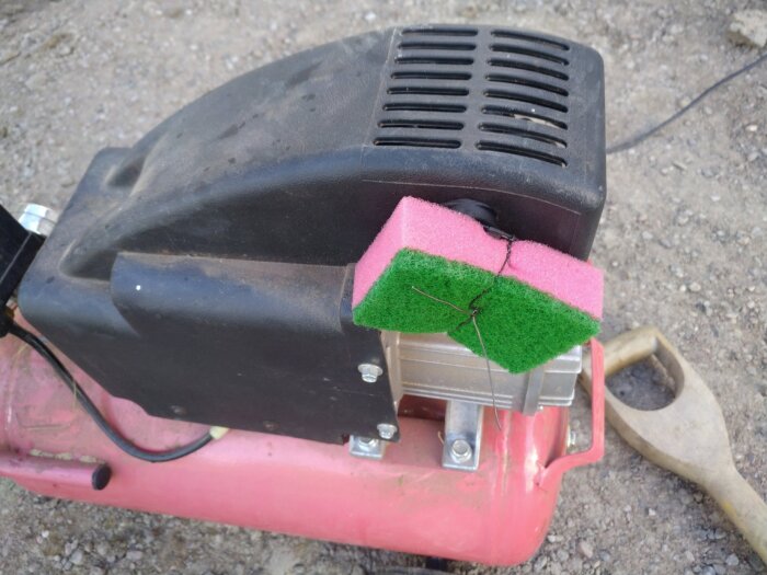 Luftkompressor med rosa och grön rengöringssvamp fastbunden vid ventilen med svart tråd. Bakgrunden är grusig mark med en spade synlig.