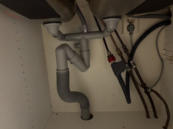 Köksunderskåp med synliga avloppsrör, slangar och eluttag, ingen lokal avstängningsventil synlig.