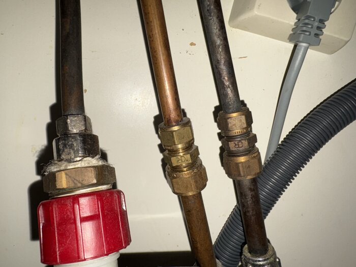 Vattenrör under diskbänk med kopplingar och rördelar, inklusive ett rött plastreglage och grå kabel.