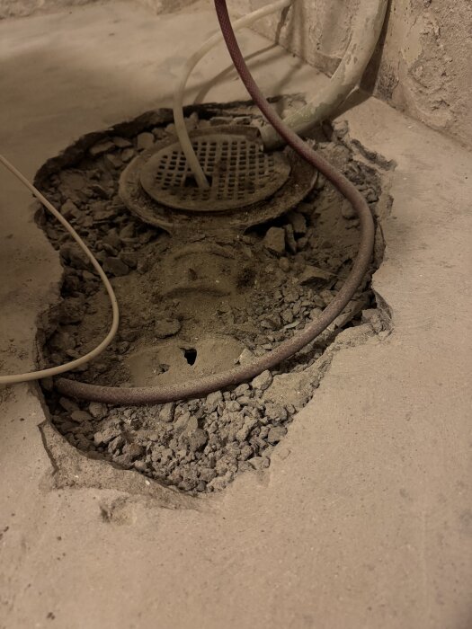 Golv med uppbrutet betong runt ett avloppsgaller och synliga rör i en källare under renovering.