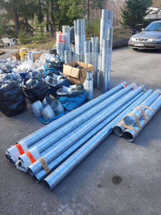 Bild av flera staplade ventilationsrör och tillbehör på en uppfart, med en personbil i bakgrunden och skog runt omkring.