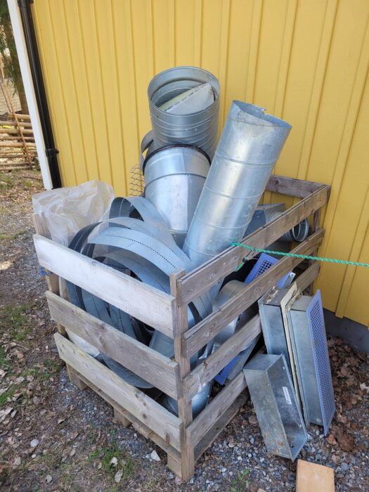 En träinklädd pall fylld med galvaniserade ventilationsrör och delar som montageband, böjningar och kanaler, vid sidan av en gul vägg.