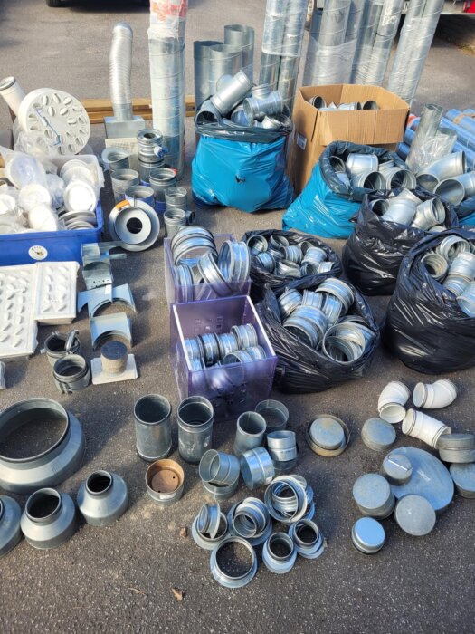 Ventilationsmaterial till salu: olika rör, delar och tillbehör i metall, uppställda på marken i lådor och säckar, redo för upphämtning.