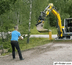 Person och grävmaskin spelar badminton med en vit boll på en grusväg omgiven av träd och grönska.