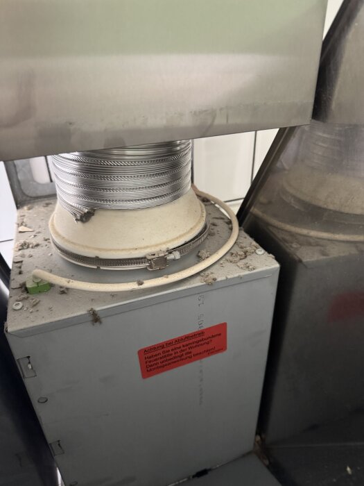 Bild av köksfläktens insida med synliga rör och en fästklämma, märkning på enhetens yta. Smuts och damm syns på ytan.