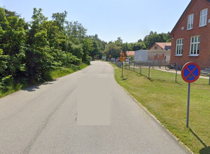Ingångsväg till skolområde med varningsskyltar för barn. Cykelställ och parkeringsförbudsskylt synliga vid trottoarkanten. Asfalterad väg svänger.