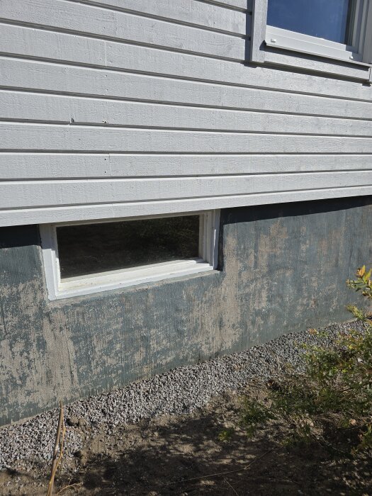 Sliten husgrund med flagnande färg och delvis exponerad puts under ett fönster, bredvid grusspridning och buskar, under solig dag.