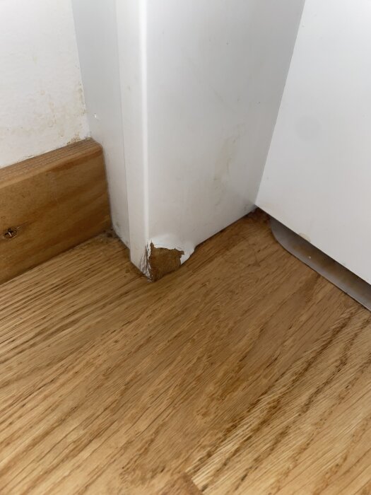 Vattenskada på vit list vid golv, med svällt och färg som släppt i hörnet, orsakat av vatten från kaffekokare.