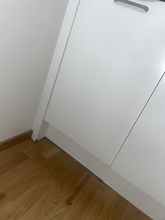 Vit kökslist med vattenskada i hörnet, där färgen släppt och listen svällt. Träparkettgolv under listen.