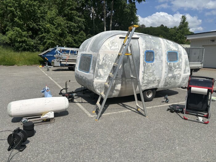 Husvagn under renovering på parkeringsplats, förberedd för målning med stege, plasttäckta fönster, och målningsutrustning bredvid.