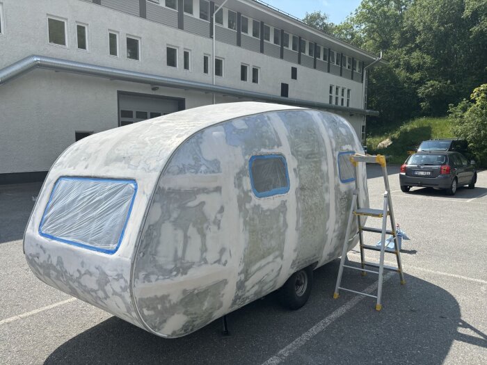 Husvagn utomhus under renovering, fönster täckta med plast. Stege lutar mot sidan, redo för målning.