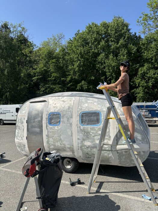 En person står på en stege och förbereder en vagn för målning. Vagnen är slipad och fönstren täckta med plast. Soligt väder och grönska i bakgrunden.
