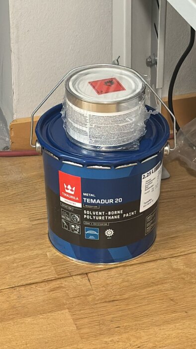 En burk med blå etikett innehållandes Temadur 20, en lösningsmedelsbaserad polyuretanfärg, står på ett golv vid en vit vägg.