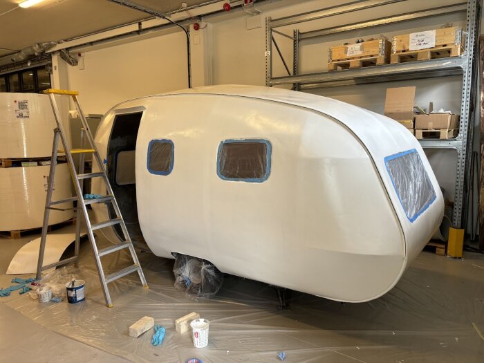 En nymålad äggskalsvit vagn med maskerade fönster står i en verkstad. En stege och målarutrustning syns bredvid.