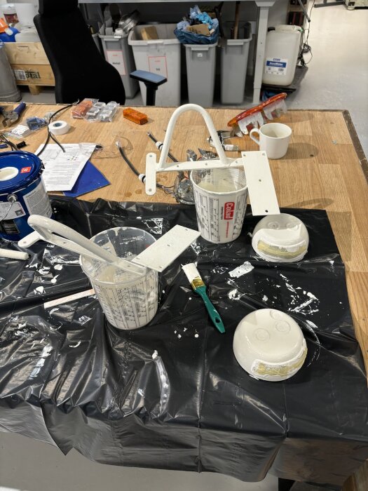 Verkstadsyta med målarverktyg, vitt färgmätglas, målade handtag och baklyktor, täckt med svart plastduk för skydd.