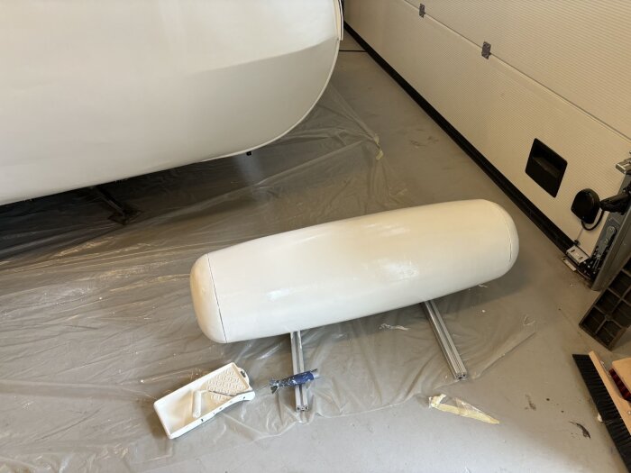 Vitmålad vagn och komponenter på plastskyddat golv i garage; färgroller och bricka synliga, delen målad med äggskalsvit polyuretanfärg.