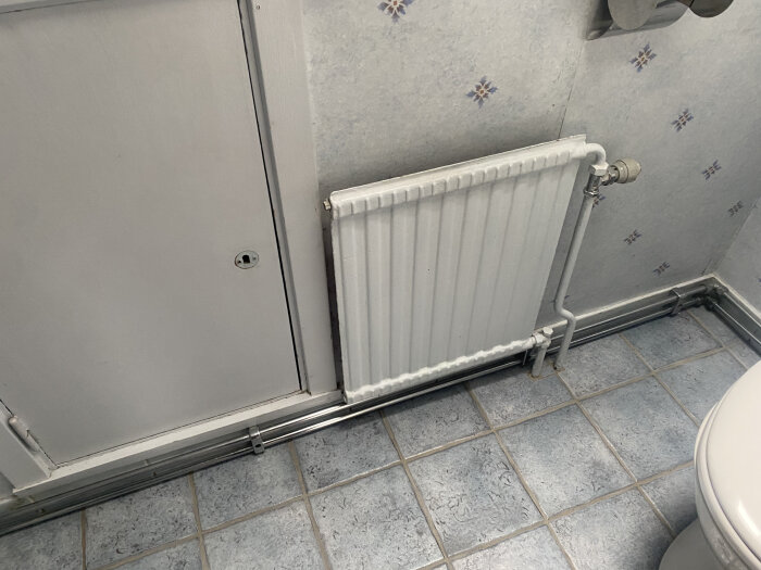 Radiator och röranslutningar i ett badrum med blåa kakelplattor och blomstrande väggmönster.