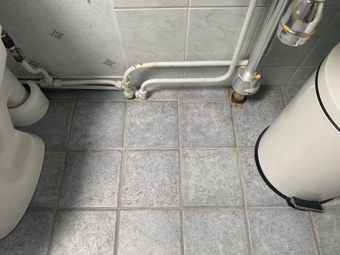 Rörstick upp ur badrumsgolvet med kakelplattor och en vit hink synlig bredvid installationen.