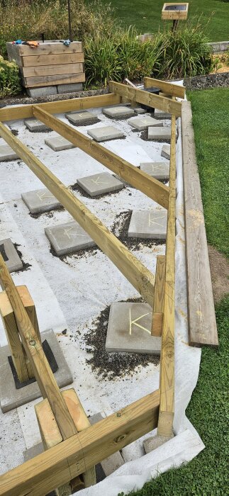 Träram för altan/trall under uppbyggnad, placerad på betongplattor. Verktygslåda och handskar synliga i bakgrunden. Grönska runtom.