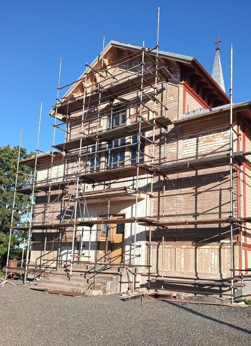 Trähus med byggnadsställningar och färg bortskrapad från väggarna under renovering i solsken.