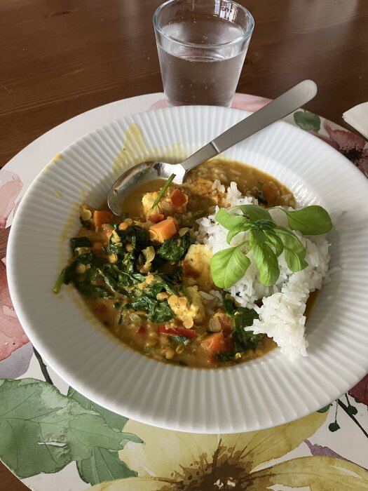 Daal med spenat, morötter och paneer serveras med ris, dekorerat med basilika. Glas med vatten i bakgrunden på ett blommigt underlägg.