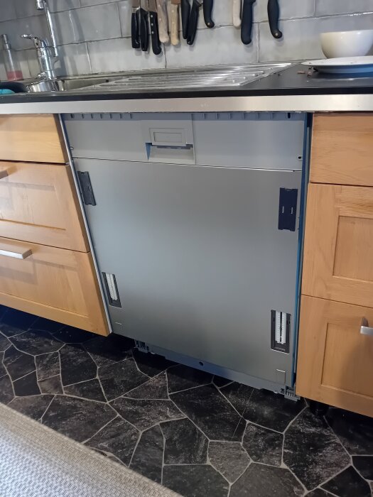 Omonterad diskmaskin under köksbänk mellan skåp, med verktyg på bänken och svartvitt stengolv.