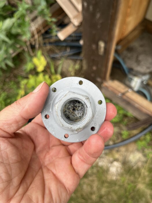 En hand håller en rund, silverfärgad metallfläns med sex hål, i en trädgårdsmiljö med trä och växter i bakgrunden.