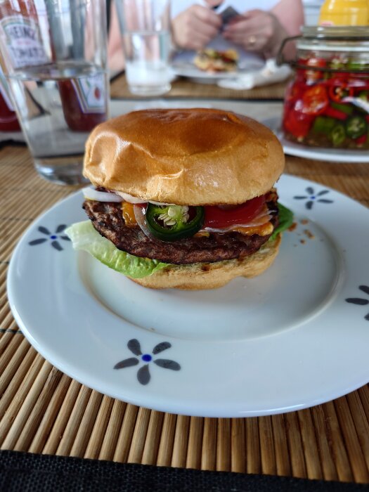 Hamburgare med sallad, tomat, picklad chili och dressing på tallrik, omgiven av glas och syltburk i bakgrunden.