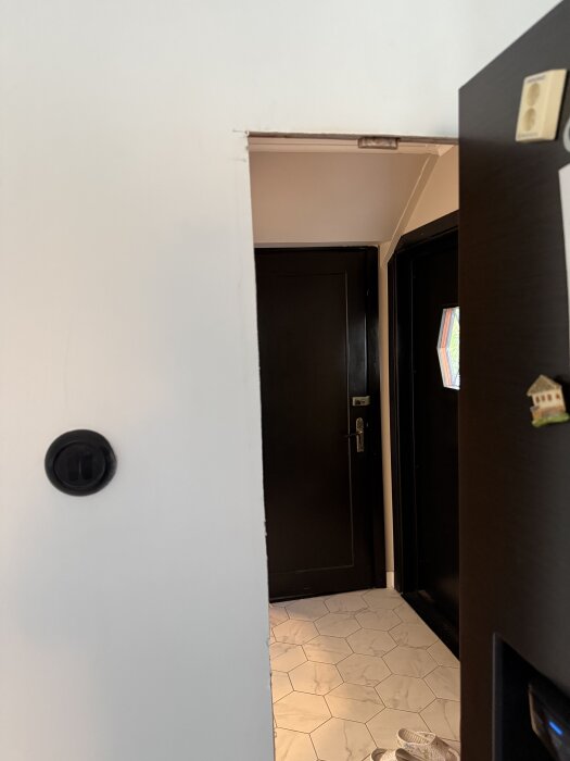 En öppning i en vit vägg in mot en hall med mörka dörrar och ljus hexagonformad golvplatta är synlig.