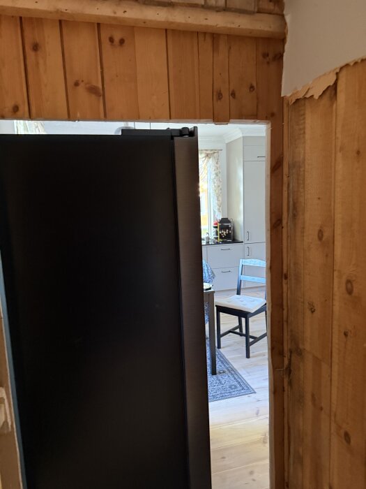 Side by side-kylskåp delvis synligt genom öppning i trävägg, med köksmöbler i bakgrunden. Projekt att bygga in kylskåpet i väggen.