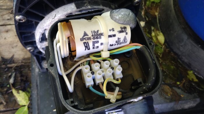 Kondensator på 10uF inne i en skadad vattenpump, omgiven av sladdar och anslutningar, med synliga skador i plasten.