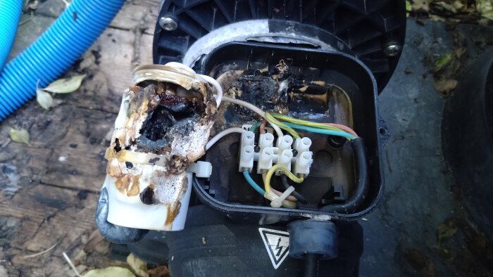 Trasig och bränd kondensator i pump, med exponerade kablar och smältskador, liggande på en grov trägolvsyta med blå slang i bakgrunden.