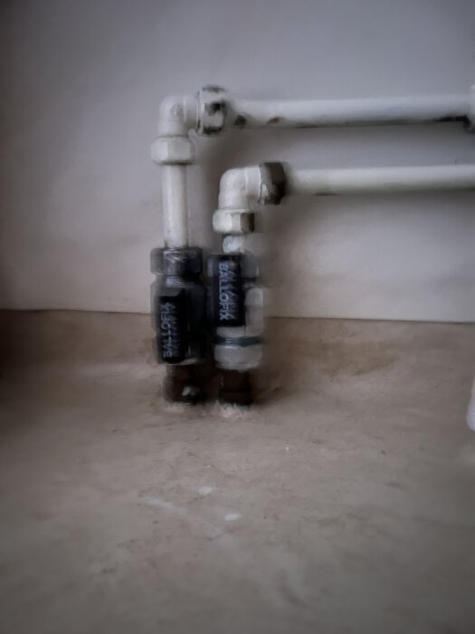 Vattenrör som kommer upp ur ett badrumsgolv med kopplingar och ventiler synliga mot en vit vägg.