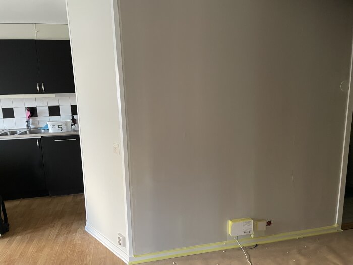 Vägg med ofullständigt spacklat område upp till taket i ett rum. Del av kök med mörka skåp synligt i bakgrunden.